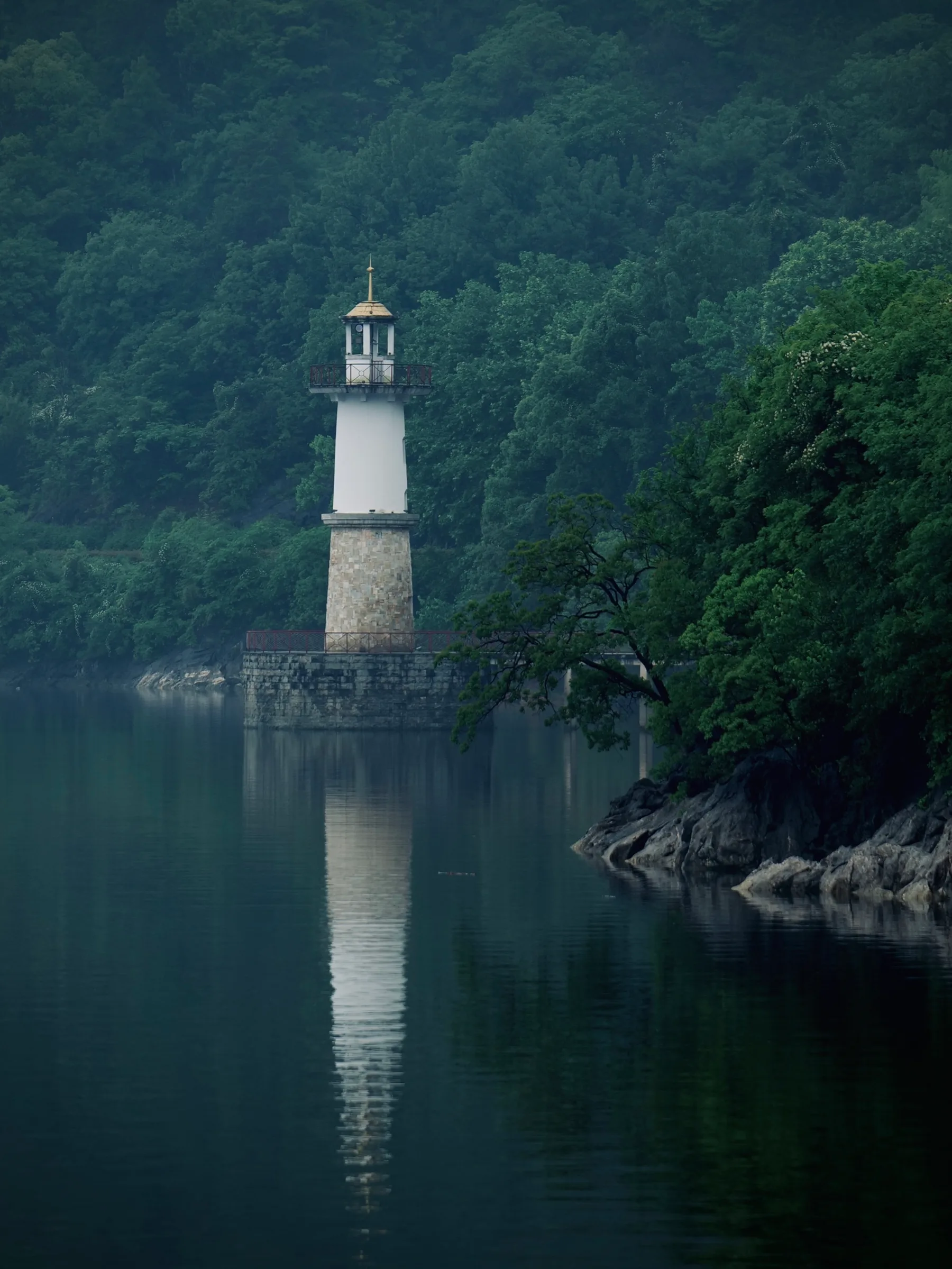 A white lighthouse reflected in a calm lake beneath dense green forest
