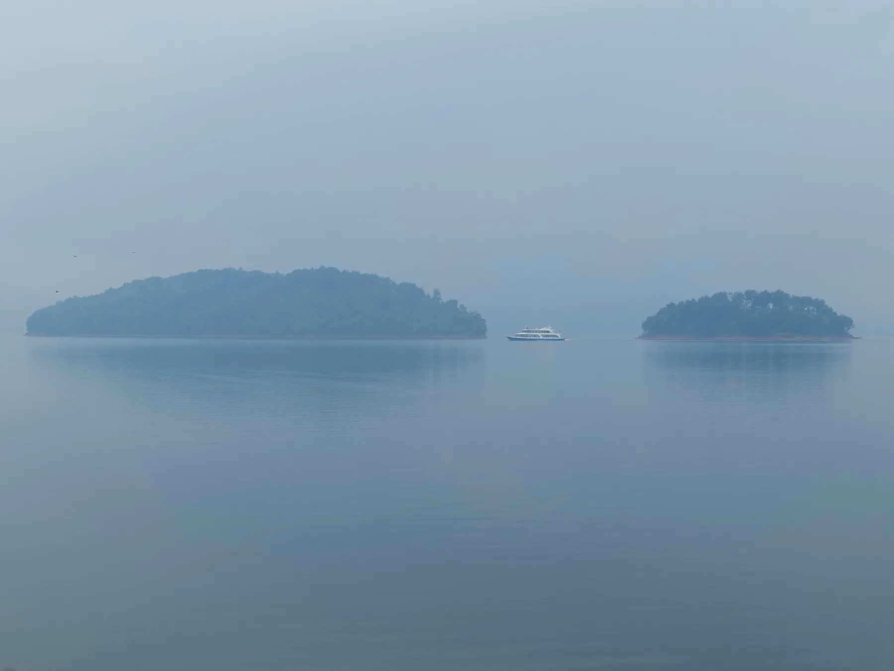 A small ferry passing between two wooded islands on a misty blue lake