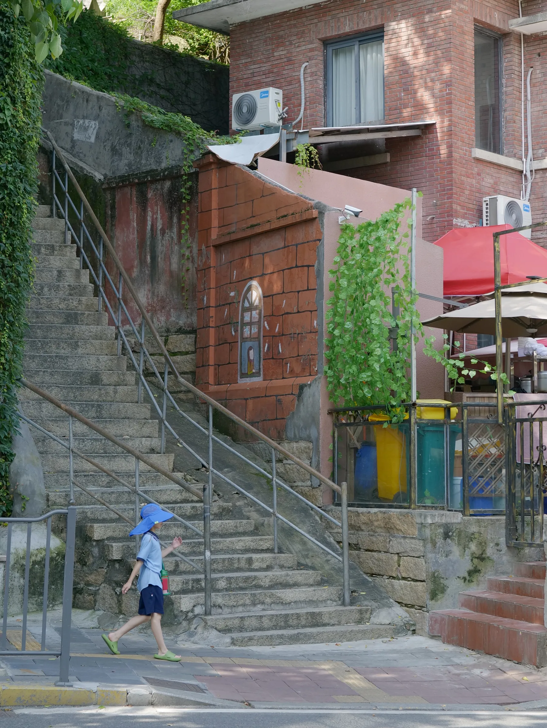 A child in a blue sun hat walking past steep stone steps and red brick buildings covered with green vines