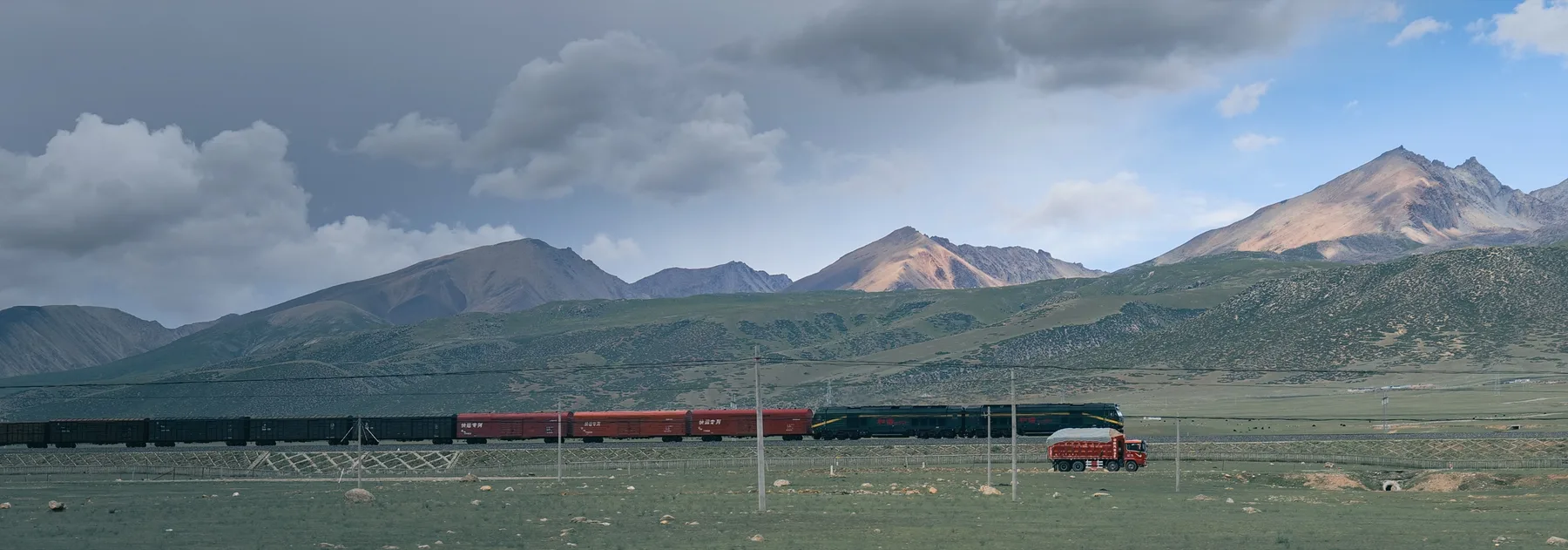 A red train crossing an open plain beneath dramatic mountain clouds