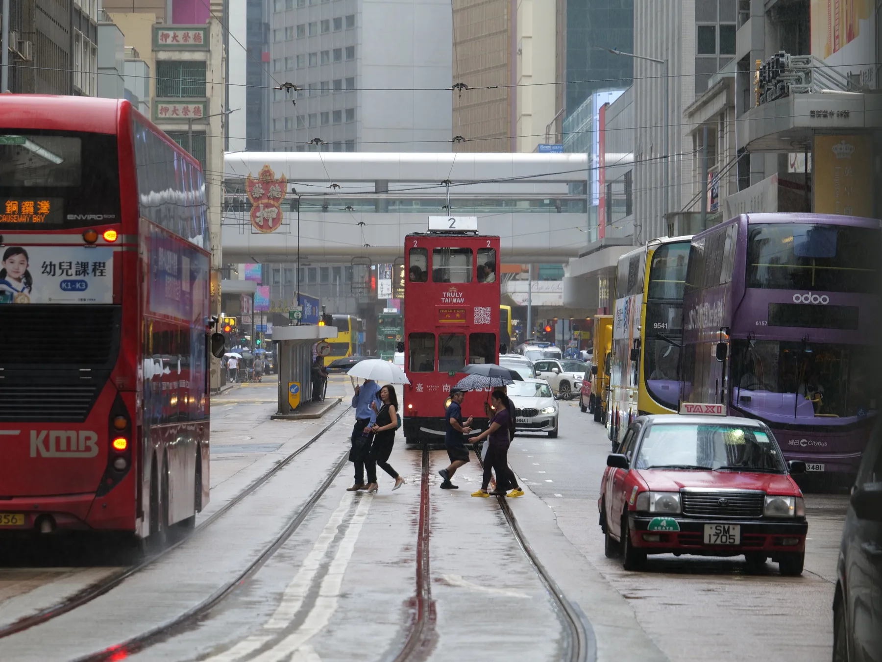 Pedestrians crossing wet tram tracks among buses, a red tram, and taxis in Hong Kong